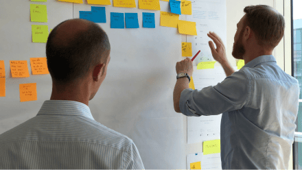 Two people post sticky notes to a white board.